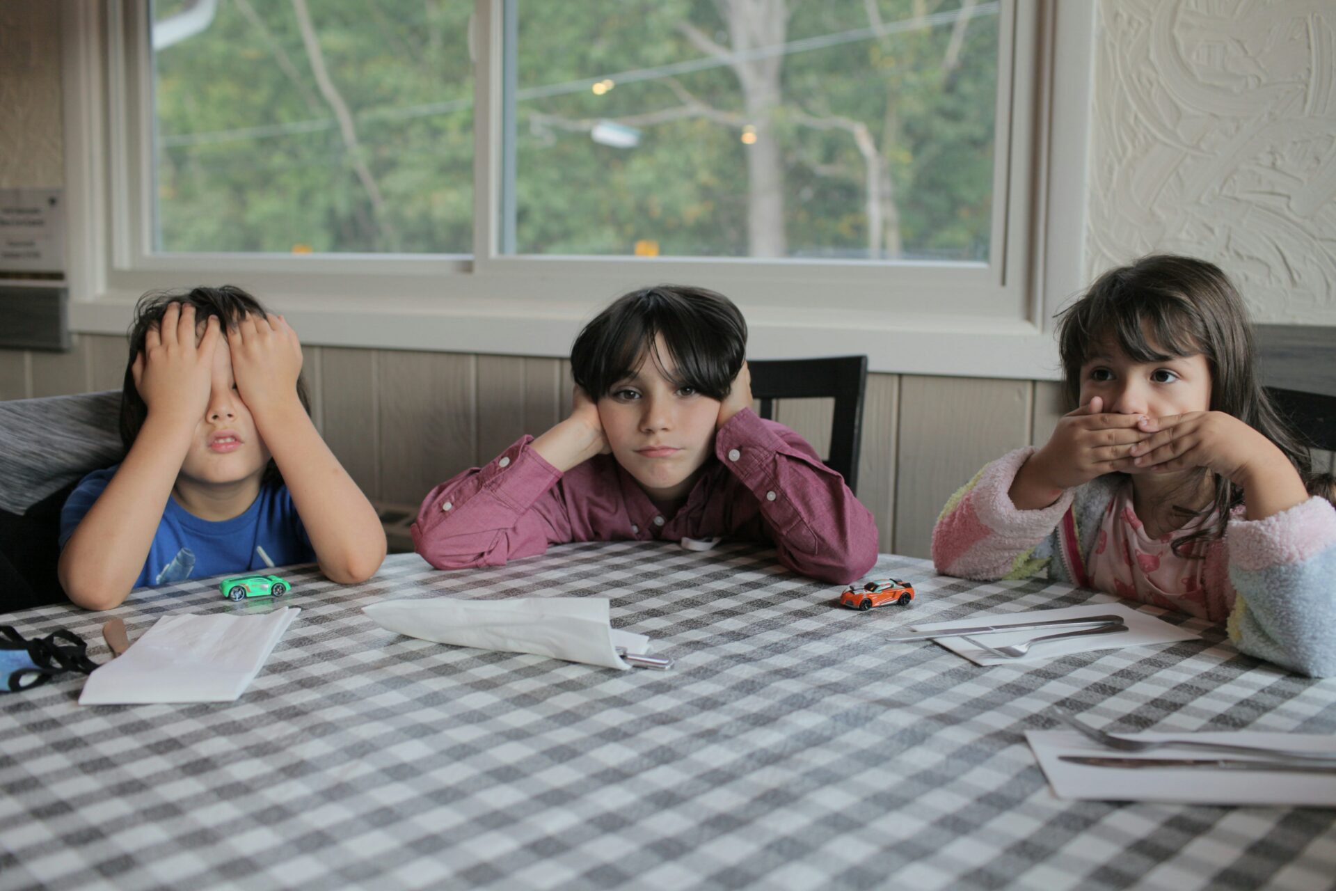 Three kids sit next to each other, one covering their eyes, one covering their ears, one covering their mouth.