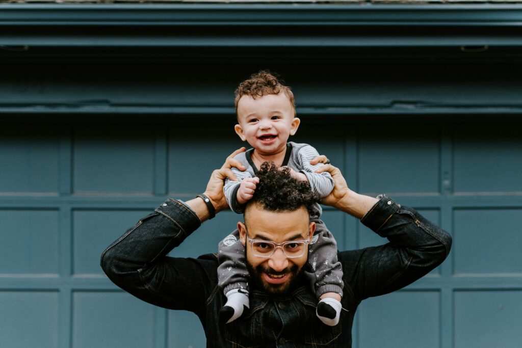 A baby boy sits on his dad's shoulders and laughs.