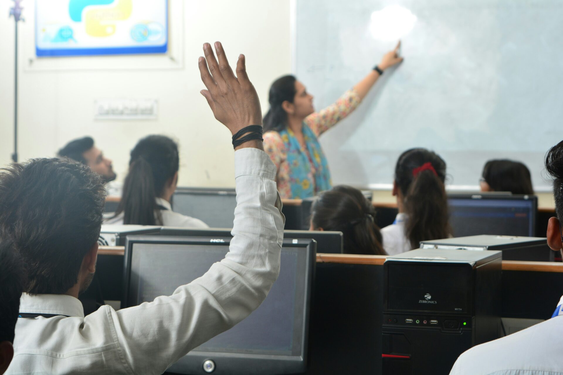 A student raises a hand to ask a question while an instructor teaches at the front of a classroom.