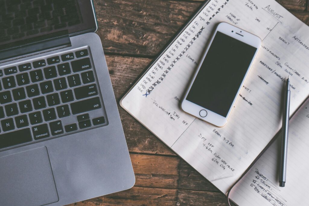 A top-down view of a laptop, notebook open to a monthly calendar, and a phone.