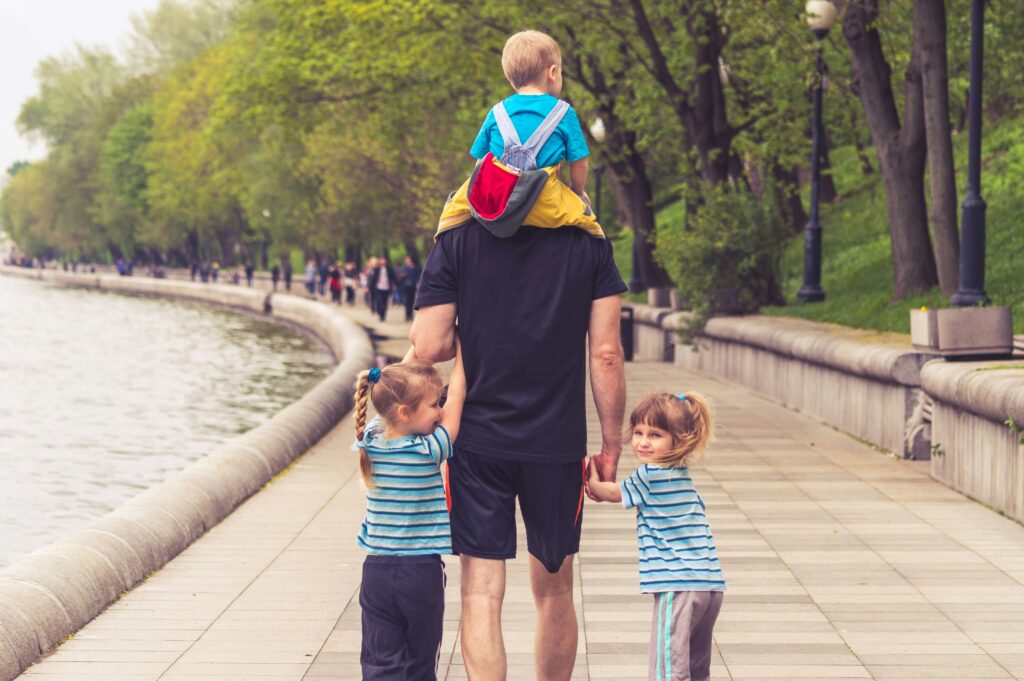 A dad walks with his three kids: one riding on his shoulders, the other two holding his hands.